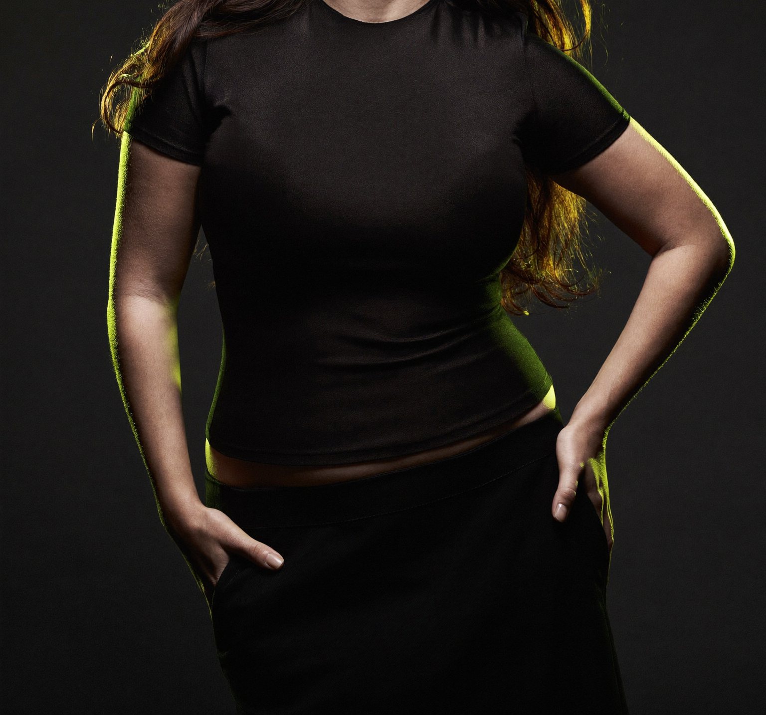 A confident adult standing tall in a fitted short-sleeve top after completing a major weight-loss journey, fully clothed, matte-black editorial setting